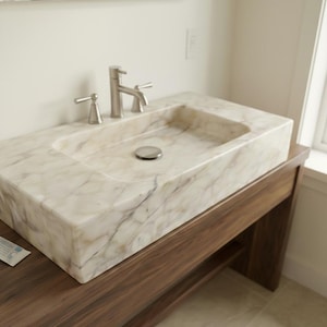 Puede incluir: Un lavabo rectangular de mármol con una pila integrada y un grifo de níquel cepillado. El lavabo está colocado sobre un mueble de baño de madera oscura con un estante inferior. El mármol tiene un veteado crema y marrón.