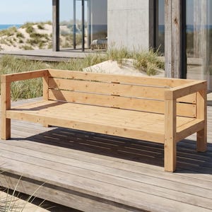 Op de afbeelding: Een lichtgekleurde houten buitenbank met een rechthoekig frame en armleuningen. De bank heeft een lattenrugleuning en zitting en is geconstrueerd met zichtbare schroeven. De bank staat op een houten terras, met uitzicht op de oceaan en zandduinen op de achtergrond.