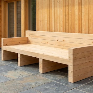 Può includere: Una panca in legno di colore chiaro con schienale e braccioli. La panca è costruita con assi orizzontali e poggia su quattro supporti rettangolari. La panca è all'aperto, con un edificio in legno e un patio in pietra sullo sfondo.