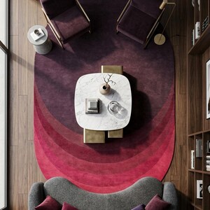 May include: An oval area rug featuring a gradient of concentric rings in shades of deep purple, magenta, and bright red. The rug is positioned beneath a white marble coffee table, two dark purple armchairs, and a curved gray sofa with four pillows.