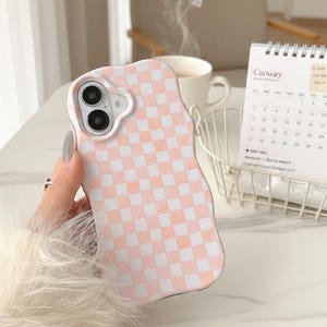 May include: A pink and white checkered phone case with a wavy edge. The case has a camera cutout and is held by a person. A calendar and a cup of coffee are in the background.