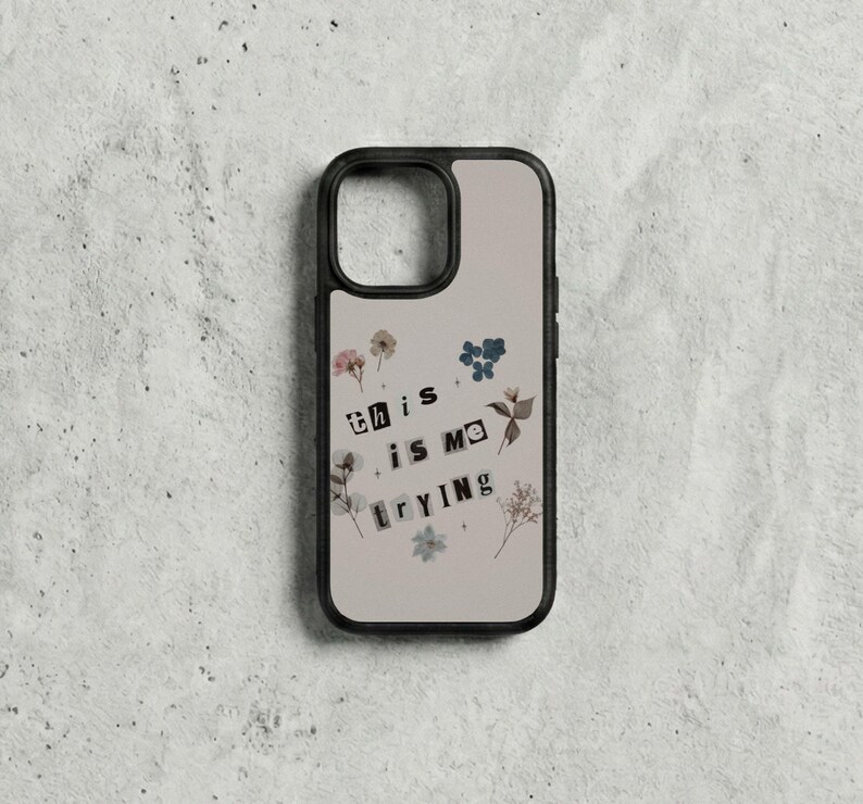 May include: A phone case with a black border and a beige background. The case features the text "this is me trying" in a collage style, with small floral and botanical elements.