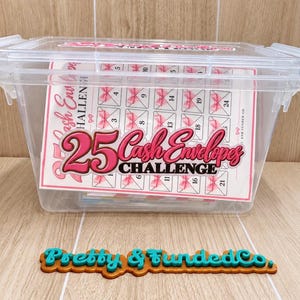 May include: A clear plastic storage box containing a 25 Cash Envelopes Challenge tracker sheet with pink bow graphics and numbered squares. In front of the box sits a brown and teal sign with the text Pretty & Funded Co. in a script font.