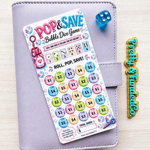 May include: A Pop & Save Bubble Dice Game savings tracker card with colorful bubble illustrations, dollar amounts, and a dice key. Includes a blue translucent die and a vertical wooden cutout reading Pretty & Funded Co. on a lavender binder.