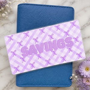 Könnte beinhalten: Ein blaues Notizbuch mit einer transparenten Kunststoffauflage. Die Auflage zeigt das Wort "SAVINGS" in lilafarbenen Buchstaben, vor einem hellvioletten Vichy-Muster mit Schleifenakzenten. Blumenelemente umgeben das Notizbuch.