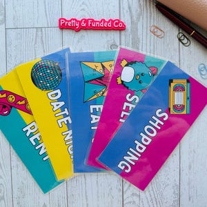 Könnte beinhalten: Eine Sammlung farbenfroher Budgetumschläge mit verschiedenen Designs und Texten. Die Umschläge sind mit "RENT", "DATE NIGHT", "EAT OUT", "SELL" und "SHOPPING" beschriftet. Ein Stift, Büroklammern und ein kleines rosa Täschchen sind ebenfalls zu sehen.