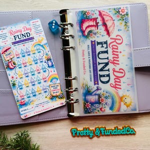 May include: A light purple six-ring binder containing a Rainy Day Fund savings tracker card with raindrop icons, a translucent dashboard with a pink umbrella and boots illustration, a blue die, and a Pretty & Funded Co. logo cutout.