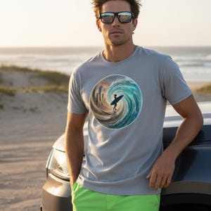 Puede incluir: Camiseta gris claro con un gráfico circular de un surfista surfeando una ola. El surfista está silueteado contra una escena oceánica arremolinada. La persona lleva pantalones cortos verdes y gafas de sol.