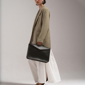 May include: A person wearing a tan blazer, white dress, and black shoes, holding a black leather briefcase. The outfit is simple and elegant, with a focus on clean lines and neutral colors. The briefcase has a flap closure.