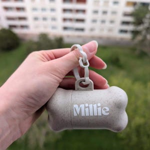 Peut inclure: Distributeur de sacs à déjections canines beige clair en forme d'os, avec le nom "Millie" imprimé en blanc. Le distributeur est maintenu par un clip en plastique blanc et est conçu pour être attaché à une laisse. L'arrière-plan est flou.