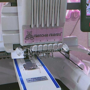 May include: A white and gray embroidery machine with the text "FRENCHIE FRIENDS" on the front. The machine has multiple needles and a blue and white support bar. The machine is designed for fabric embroidery.