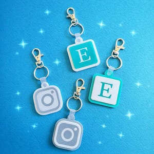May include: Four NFC keychains with silver rings and gold clasps are displayed on a blue background. Two keychains have a white square with a teal border and the letter "E". The other two have a gray Instagram logo.