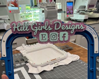 Puede incluir: Un bastidor de bordado azul con un letrero personalizado que muestra el texto Hill Girls Designs en letras verde claro sobre un fondo de purpurina morada, junto a tres iconos cuadrados verde claro con las letras E, un logo de Instagram y una f.