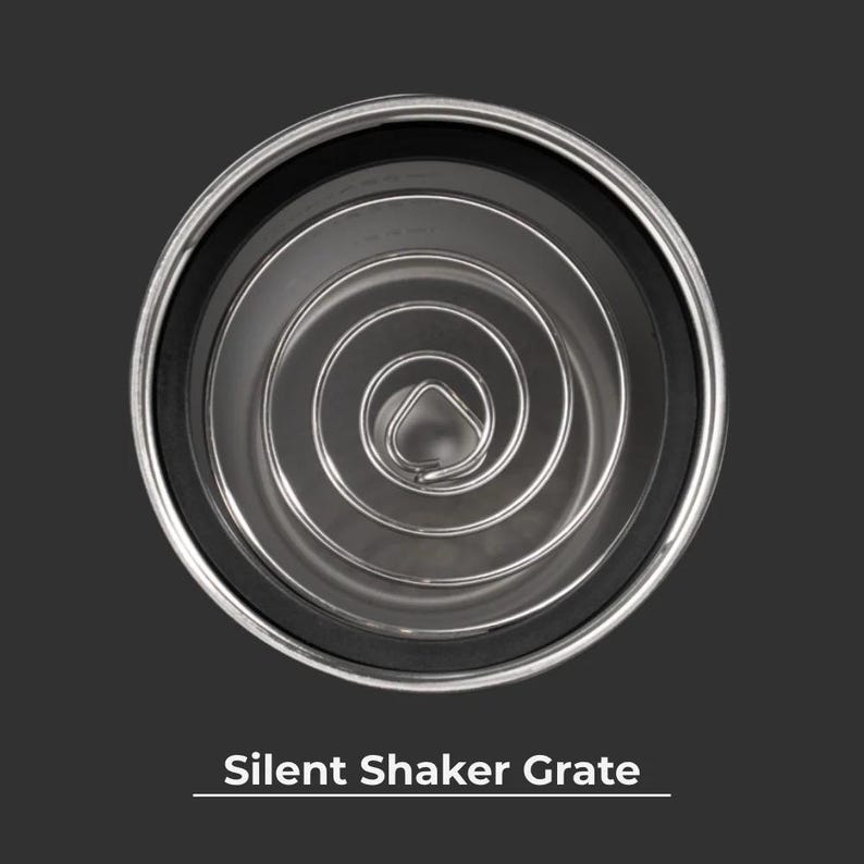 May include: A top-down view of a stainless steel silent shaker grate. The grate features a coiled wire design within a black rubber ring and a silver rim. The text "Silent Shaker Grate" is displayed at the bottom of the image.
