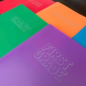 May include: A collection of colorful folders in red, green, orange, purple, and blue. The red folder is labeled ENGLISH, the green folder is labeled GRANT, the orange folder is labeled HISTORY, and the purple folder is labeled FIRST GRADE.