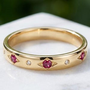 Op de afbeelding: Een gouden bandring met een reeks afwisselende robijn- en diamantaccenten. De robijnen zijn gezet in een sterpatroon en de diamanten zijn klein en rond. De ring wordt gepresenteerd op een wit oppervlak.