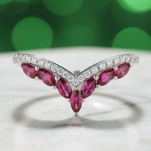 Puede incluir: Un anillo plateado con un diseño en forma de V. El anillo presenta una fila de pequeñas piedras transparentes en la parte superior y una serie de piedras preciosas rosas oscuras con corte marquesa en el centro. El anillo está sobre una superficie blanca con un fondo verde borroso.