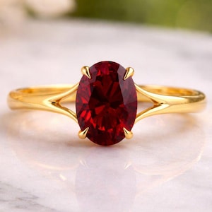 May include: A gold ring featuring a large, oval-cut, deep red gemstone. The ring's band splits into a delicate design on either side of the stone, creating an elegant and eye-catching piece of jewelry.
