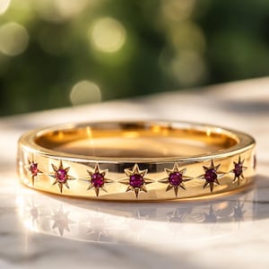 May include: A gold ring with a band featuring starburst designs, each containing a small, round, red gemstone. The ring is set on a marble surface, reflecting the sunlight. The ring's design is elegant and eye-catching.