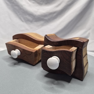 May include: Two wooden boxes with drawers. One box is open, revealing the interior. The boxes are made of various wood tones, including light and dark browns. Each box has a white, round knob on the drawer.