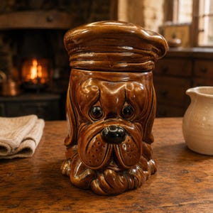 May include: A brown ceramic cookie jar shaped like a dog wearing a beret. The dog has large, dark, glossy eyes, a black nose, and textured jowls. The lid is formed by the dog's hat, which sits atop the head. The finish is smooth and glazed.