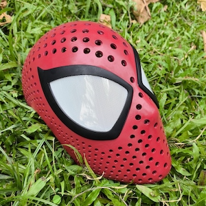 May include: A red mask featuring a perforated surface with numerous small circular holes throughout the exterior. The mask includes two large, almond-shaped eye openings outlined in black, covered with a fine white mesh screen material.