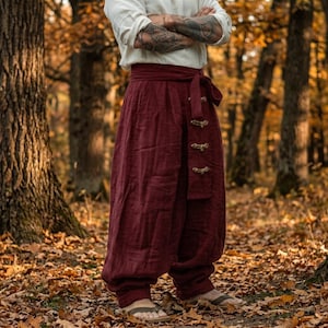 Peut inclure: Pantalon ample rouge foncé doté d'une large ceinture avec un lien de serrage latéral. Le panneau avant présente une rangée verticale de trois fermetures décoratives de type bascule. Le vêtement est associé à une chemise blanche unie.