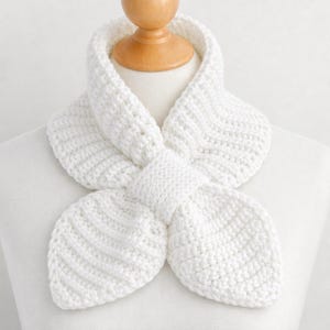 Könnte beinhalten: Ein weißer, handgehäkelter Schal, der auf einer Schaufensterpuppe präsentiert wird. Der Schal hat ein einzigartiges Design mit einem geknoteten Mittelteil und ausgestellten Enden. Das strukturierte Strickmuster erzeugt ein weiches, gemütliches Aussehen. Der Schal ist ein vielseitiges Accessoire.