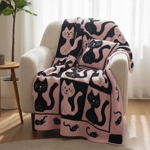 May include: A pink and black crocheted blanket and pillow set featuring a repeating cat design. The blanket is draped over a cream-colored armchair. The cat design is black on a pink background.