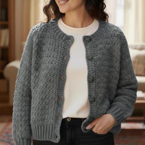 Może przedstawiać: Szary, zapinany na guziki kardigan z szydełka z okrągłym dekoltem i długimi rękawami. Kardigan ma wzorzysty ścieg i guziki o wyglądzie drewna. Osoba ma na sobie biały t-shirt i czarne dżinsy.