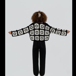 May include: A black and white crochet cropped jacket with a square pattern. The jacket is worn with black jeans. The model has curly hair and a white hair clip.