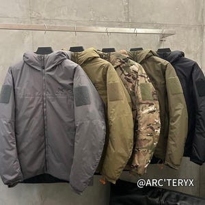 Puede incluir: Una fila de chaquetas con capucha en varios colores, incluyendo gris, verde oliva, camuflaje y negro, colgadas de una barra metálica. Cada chaqueta tiene un parche en la parte superior del brazo y cierre de cremallera. Se ve la marca "ARC'TERYX".