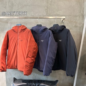 Puede incluir: Tres chaquetas con capucha en óxido, azul marino y negro cuelgan de una barra metálica. Cada chaqueta tiene una cremallera frontal y un pequeño logotipo en el pecho. Las chaquetas se exhiben contra una pared gris neutra. La marca "@ARC'TERYX" es visible.