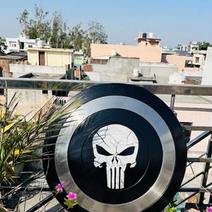 May include: A black and silver shield with a white skull emblem. The shield is circular, with a black centre, a silver ring, and a black outer edge. The skull design has a cracked effect.