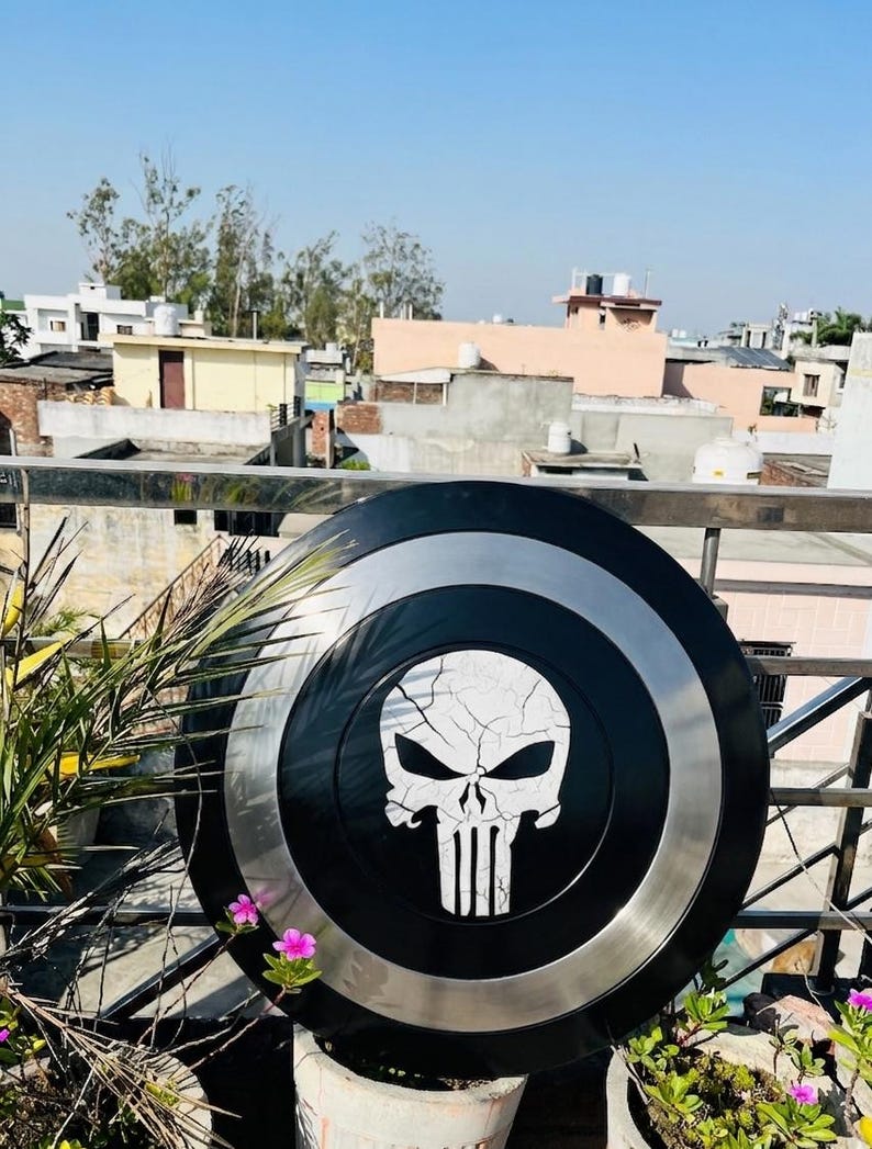 May include: A black and silver shield with a white skull emblem. The shield is circular, with a black centre, a silver ring, and a black outer edge. The skull design has a cracked effect.