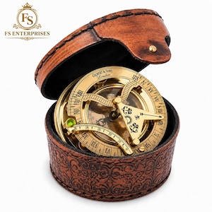 May include: A brass sundial compass with Roman numerals and a central gnomon, engraved with the text Gilbert & Sons London. The instrument is housed in a round, brown, embossed leather case with a hinged lid and visible stitching details.
