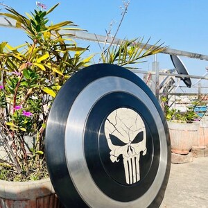 May include: A circular shield in black and silver, emblazoned with a white skull motif. The shield has a silver band around the central black area. The skull design has a cracked effect. The shield is propped up near a plant pot.