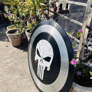 May include: A black and silver shield with a white skull emblem. The shield is circular, with a black border, a silver band, and a central skull design with vertical lines. The shield is displayed outside, with plants in the background.