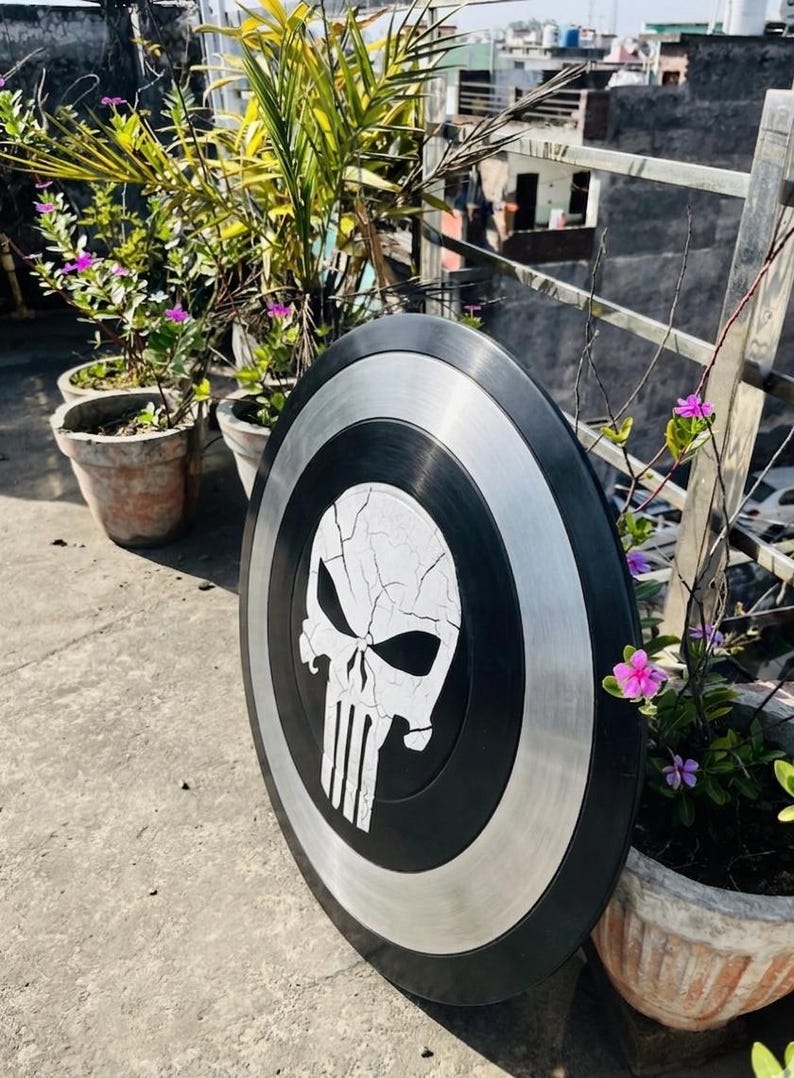 May include: A black and silver shield with a white skull emblem. The shield is circular, with a black border, a silver band, and a central skull design with vertical lines. The shield is displayed outside, with plants in the background.