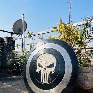 May include: A black and silver shield with a white skull emblem. The shield is circular, with a black central area and a silver border. The skull has a cracked effect, and the shield is shown outside against a blue sky.