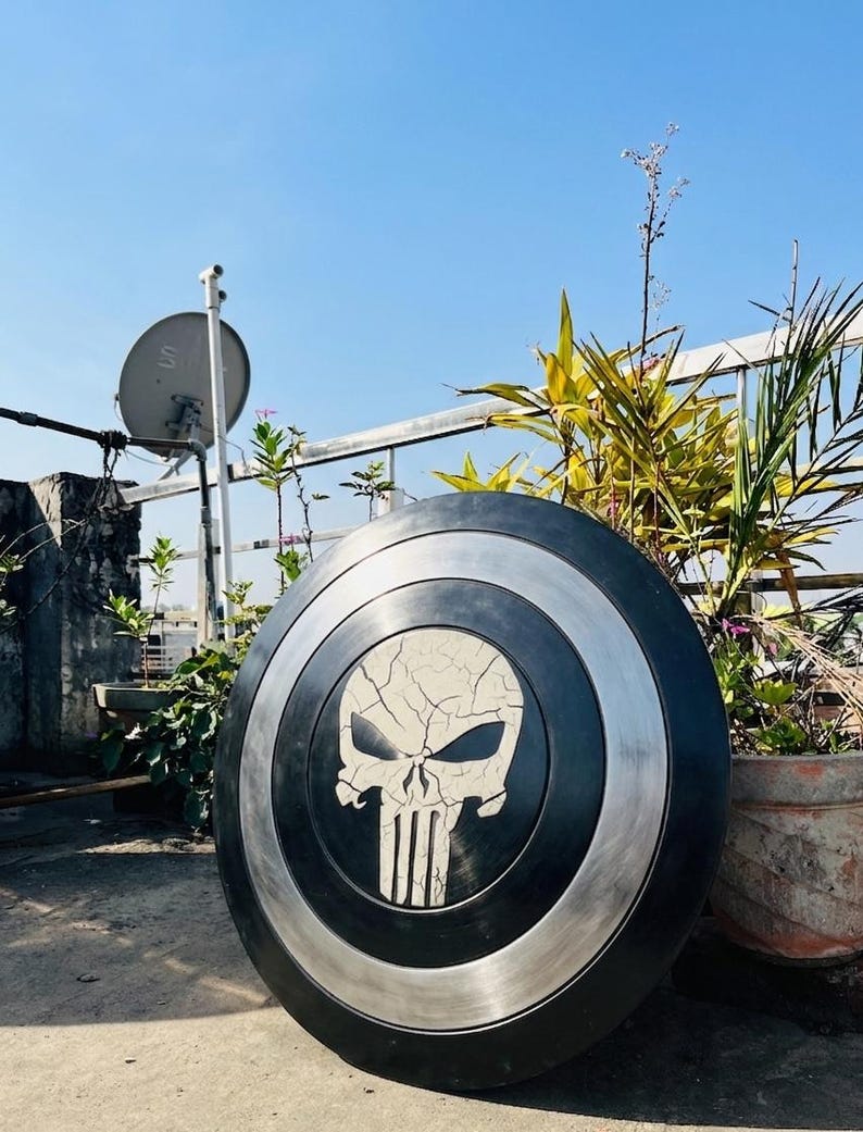 May include: A black and silver shield with a white skull emblem. The shield is circular, with a black central area and a silver border. The skull has a cracked effect, and the shield is shown outside against a blue sky.