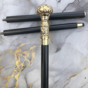 Peut inclure: Une canne noire avec une poignée dorée ornée. La poignée présente un motif floral et un dessus arrondi. Deux sections de canne noires supplémentaires sont visibles en arrière-plan.