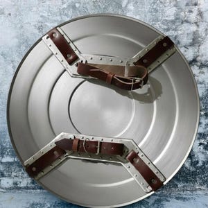 May include: A circular, silver-coloured shield with brown leather straps and metal detailing. The shield features a central circular design and is likely made of metal. The straps are riveted to the shield.
