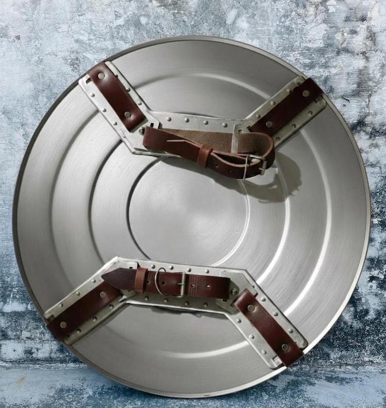 May include: A circular, silver-coloured shield with brown leather straps and metal detailing. The shield features a central circular design and is likely made of metal. The straps are riveted to the shield.