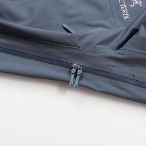 May include: Close-up of a slate blue jacket with a zipper closure. The jacket features the brand name "Arc'teryx" and the "GORE-TEX PRO" logo. The jacket has a sleek, modern design, suitable for outdoor activities.