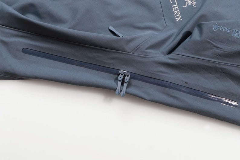 May include: Close-up of a slate blue jacket with a zipper closure. The jacket features the brand name "Arc'teryx" and the "GORE-TEX PRO" logo. The jacket has a sleek, modern design, suitable for outdoor activities.
