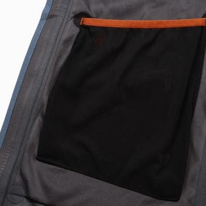 May include: Close-up of the interior of a gray jacket, showcasing a black mesh pocket with an orange trim. The jacket has a blue zipper and blue trim along the edge.