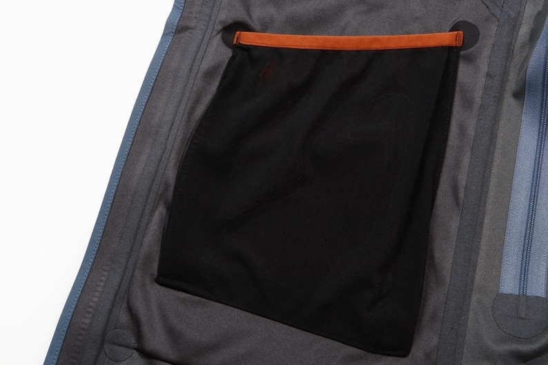 May include: Close-up of the interior of a gray jacket, showcasing a black mesh pocket with an orange trim. The jacket has a blue zipper and blue trim along the edge.