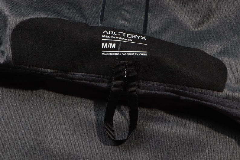 May include: Close-up of a black label from Arc'teryx, with the text "ARC'TERYX MEN'S/HOMMES M/M MADE IN CHINA/FABRIQU&Eacute; EN CHINE". The label is attached to a dark gray garment, with a black loop.