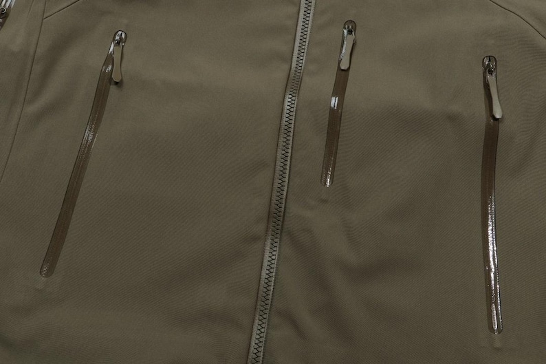 May include: Close-up of a khaki-colored jacket with three zippered pockets and a central zipper. The jacket's fabric has a subtle texture, and the zippers have silver pulls.